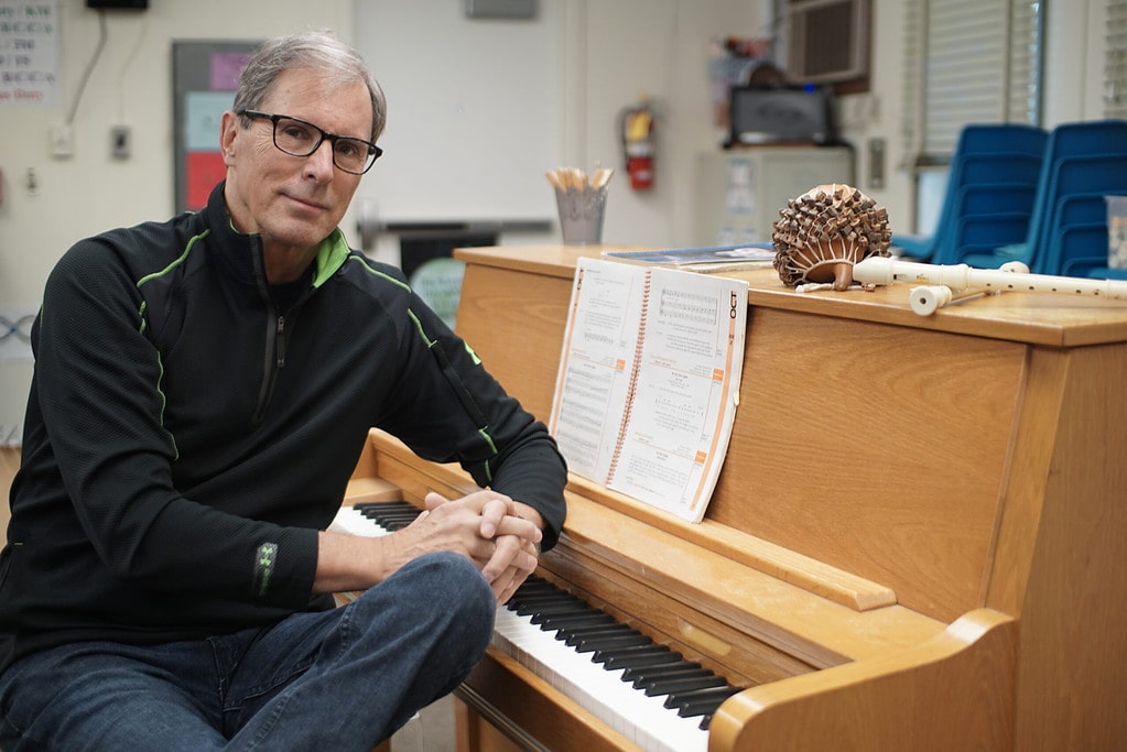 Photo of Steve Erickson posing by a piano in an elementary school.