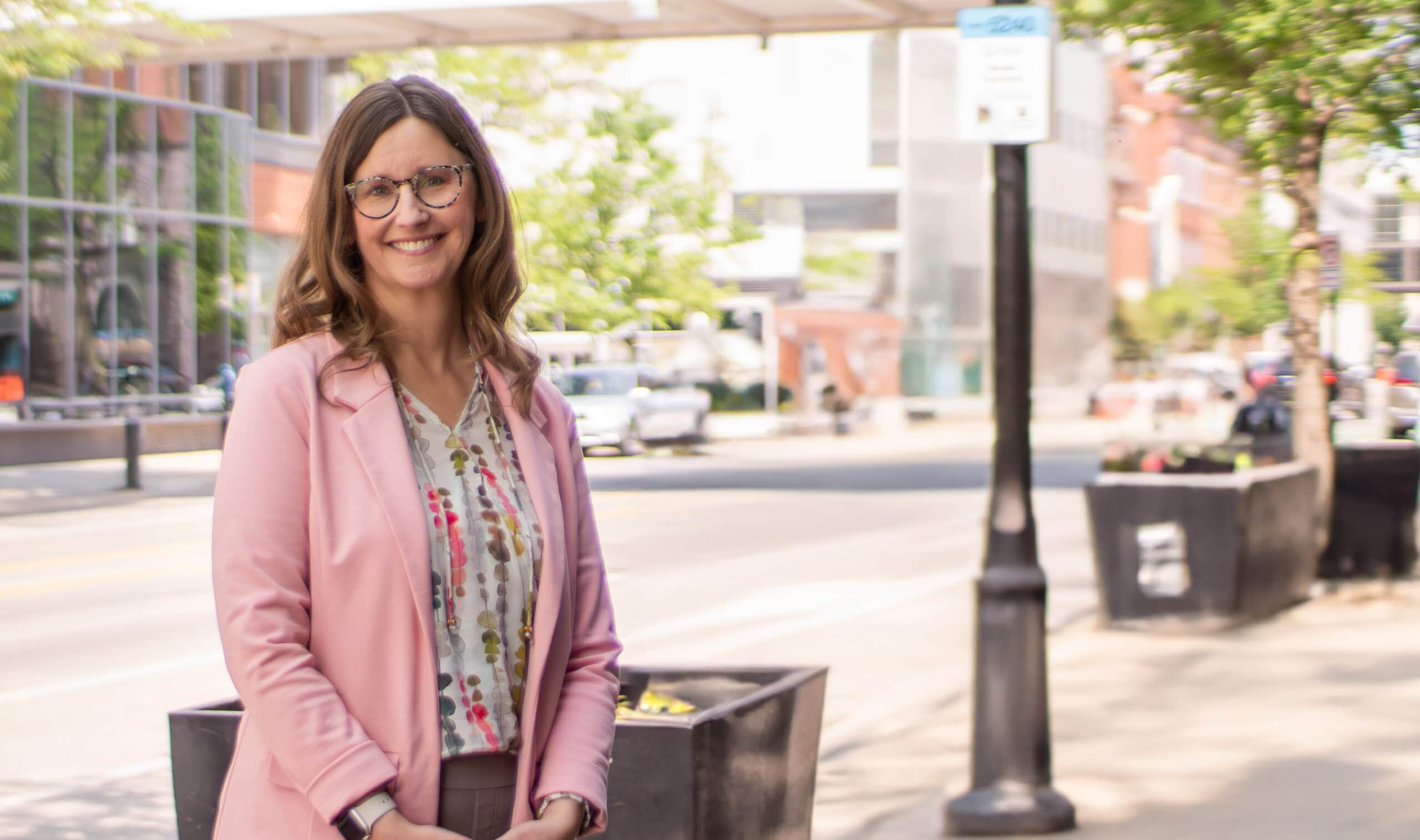 Karla Twedt-Ball smiles on a city sidewalk wearing a pink blazer.